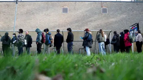 Reuters Students queue outside a light-coloured brick building. They are wearing coasts and backpacks, and some of them are wearing face masks. The image is shot from the grass and looking up.