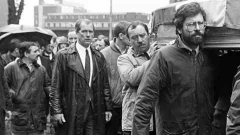 Pacemaker A black and white image of the funeral procession of Brendan Davison. To the left, almost out of frame is Scappaticci wearing a rain coat. In front is a younger Gerry Adams, carrying a coffin with a Irish tricolour on top.