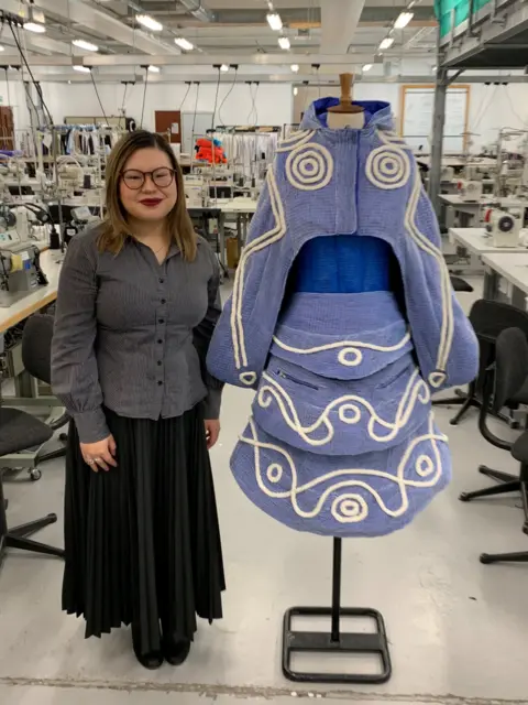 Heriot-Watt University A woman in glasses with long hair in a grey blouse and black skirt stands next to a blue outfit with white embroidery