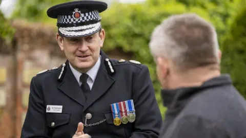 Al Underwood/Essex Police Ben-Julian Harrington is wearing a black suit with three medals on his jacket. He is also wearing a black and white police hat. He is talking to a man at an outdoor ceremony.