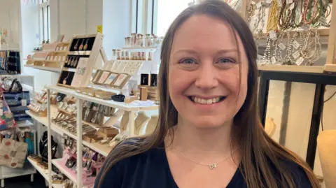 Kirsty Devine is standing in her shop in Brighouse, West Yorkshire. She has long dark hair and is smiling at the camera and is wearing a small silver necklace and black top