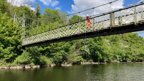 A man lowers a small bucket off a bridge above the River Wharfe at Ilkley so that he can test water pollution levels. The bucket is suspended on a rope.