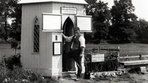 Canal & River Trust A black and white photo of a man who is a lock keeper standing outside a hut by a canal. 