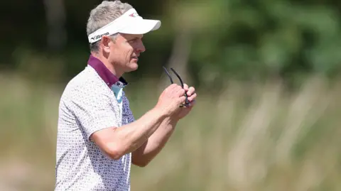 Getty Images Luke Donald photographed side on holding a pair of sunglasses in front of his face. He is wearing a white and maroon polo shirt. Donald has short greying blond hair which is visible above his white visor. 

