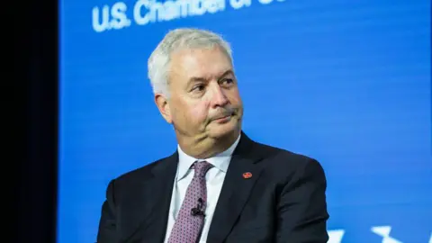 An image of Michael Rousseau sitting looking in the direction of his left. He is wearing a black suit jacket with an Air Canada logo pin, a white shirt and a patterned mauve tie. He has short grey hair.