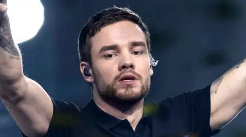 Getty Images Liam Payne, wearing a black top, holds his arms aloft while performing ion stage n 2019. He has short brown hair and a cropped brown beard and moustache