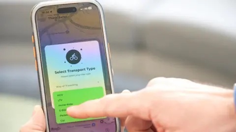 A blurred background with a phone screen in the foreground. The phone is open to a map with a selection box on it. The box says "Select Transport Type". There is a drop down menu. The visible options are HGV, LTV, Horse Rider, E-Bike, Pedestrian, Car. A finger is pointing towards the screen as if a selection is about to be made. 