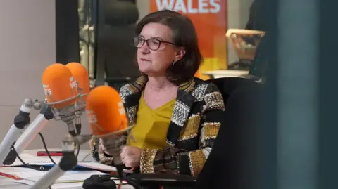 BBC The first minister Eluned Morgan sat in a radio studio wearing a multicoloured jacket and yellow top, in front of a set of BBC Radio Wales orange microphones.