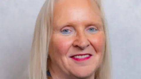 Tynwald Parliamentary portrait of Julie Edge. She has red lipstick and ash-blonde hair. She smiles in front of a grey background.