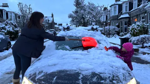 A mother and daughter clear the snow-laden windscreen of a car in the early hours. The car is on an upmarket residential street with sandstone properties, trees and bushes