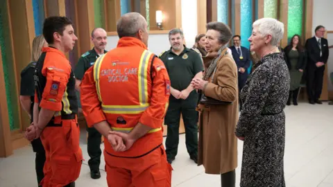 PA Media Princess Anne talks to critical care staff who are wearing orange medical scrubs in the main atrium at Southmead Hospital in Bristol. She is wearing a brown jacket and patterned scarf and other hospital staff are visible in the background