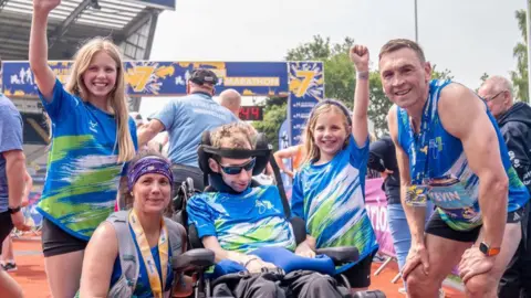 PA Media Rob Burrow alongside wife Lindsey (who ran the half marathon), daughters Macy and Maya and Kevin Sinfield who ran the full marathon all pose for a picture after the Rob Burrow Leeds Marathon