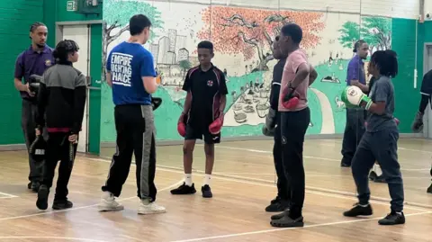 A group of young people in a sports hall. They are all wearing sports kit and boxing gloves and listening to a man wearing a blue tee shirt with Empire Fighting Chance on the back and black and grey joggers.