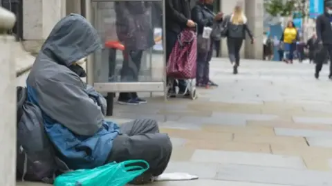 Getty Images A rough sleeper sitting by a bus stop on a busy town pavement, wearing a rain coat with the hood up and leaning against a rucksack with a plastic bag beside him