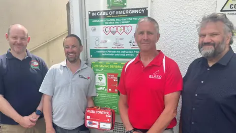 Four men standing by a public defibrillator cabinet in a row, looking at the camera. One of the men is holding a red bleed control kit in his hand.