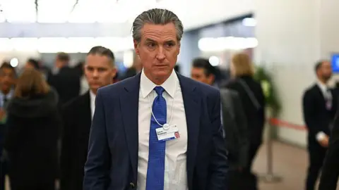California Gov Gavin Newsom is seen walking through a hallway at the Davos World Economic Forum. He has on a suit with a blue tie and has on a lanyard identifying himself. 
