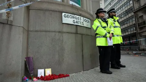 Reuters Police officers stand next to flowers and cards placed on the ground near the site of an incident where a car plowed into a crowd of Liverpool fans during a parade celebrating their side's Premier League soccer title, in central Liverpool.