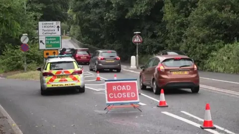 A picture of a slip road where one lane is blocked off with a red road closed sign and several traffic cones. A police car is stationed near the closure. Other vehicles are driving along a diversion. 
