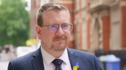 Andrew Gwynne MP is pictured in a photo taken in 2022. He is walking along a pavement with a neutral expression, wearing a dark blue suit with a pink shirt