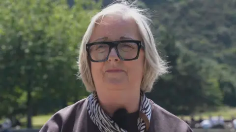 Cynthia Guthrie wears a brown top, zebra print scarf and dark glasses. She has short fair hair and is sitting outside. She looks slightly concerned 