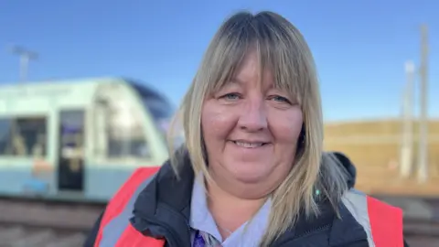 Head and shoulder image of ticket supervisor Tammy Lennon looking straight at camera. She has long blonde hair and a fringe and is wearing a pink high-vis vest over a navy jacket and pale blue shirt.
