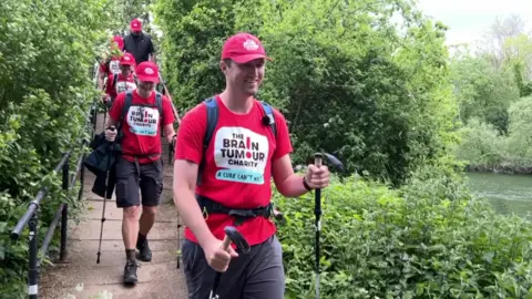 A man leading a group of people wearing red shirts for The Brain Tumour Charity. They are walking with sticks in nature.