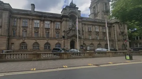 Google A building of three visible storeys. A road is in the foreground and a tree is partly visible in the top right of the photo.