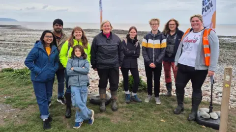 The Rockpool Project A team photo of people taking part in the Klive beach rock pool challenge. They are standing by a rocky beach, with the grey sea behind them.