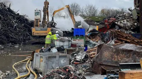 WMFS A crane is moving metal and yellow hoses are in the picture. There are also large piles of scrap metal and smoke can also be seen. Near the camera are large items of scrap metal, including boxes, sheets and blocks of metal. 