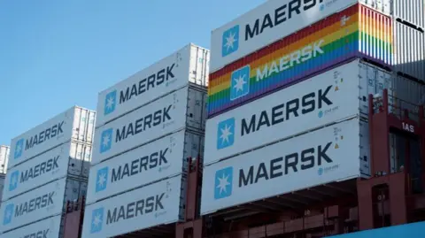 A close-up of about 12 Maersk shipping containers stacked on a ship. One of the containers is painted in horizontal rainbow strips while the rest of standard light grey  