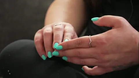 A woman holds her hands in her lap. She is wearing black and her nails are painted bright turquoise. 
