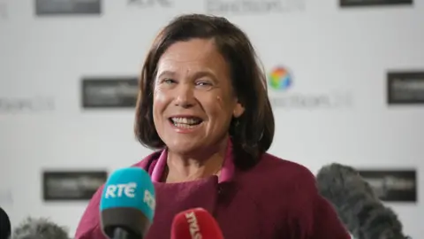 PA Media A woman with dark hair laughing in front of media microphones. She is wearing a red jumper