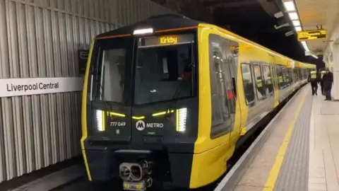 A yellow and black Merseyrail train at a platform at Liverpool Central station. The destination Kirkby is displayed in yellow at the front of the train which carries the Metro logo.