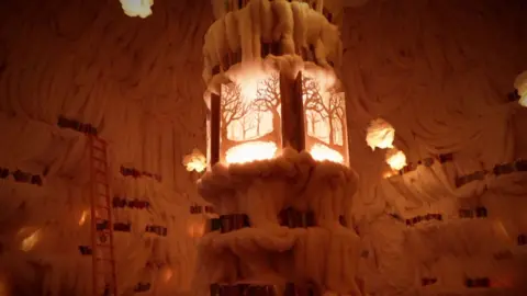 A warmly lit, intricate sculpture resembling a lantern amidst walls adorned with fluffy white formations and glowing accents.