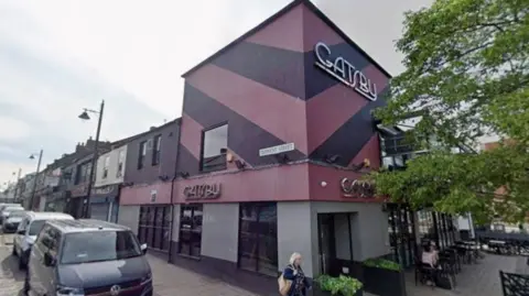 Streetview of Gatsby, a bar in Sunderland. It is a two storey block of a building painted with chevrons in two shades of purple, with trees and seats to the side of the picture. There are cars parked down the street and a woman with blonde hair is walking past. The street lamps are ornate.