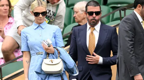 Getty Images Vestida de sangue e com uma bolsa branca pendurada no braço esquerdo, Jordyn Blum está ao lado de seu marido Dave Grohl enquanto o casal se senta no Campeonato de Tênis de Wimbledon. Grohl usa terno azul marinho, óculos escuros e gravata laranja.