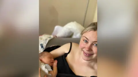 Cornelia Marland Cornelia Marland wearing a black strappy top and holding her new baby against her chest.