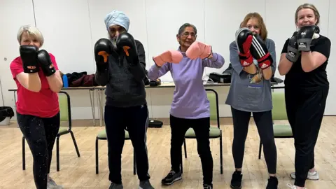 A group of women and a man with a turban hold their arms up whilst wearing boxing gloves. Various equipment and chairs can be seen in the background behind them.