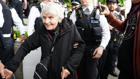 Sue Parfitt wearing a black coat and a black cross body bag. She has short white hair and is smiling and someone holds her hand and leads her through the crowd of police officers, dressed in black and white uniforms and stab proof vests.