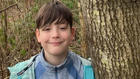 Mia Ambrose Xavier, aged 11, wears a turquoise hoodie and a zip up grey top. He has brown hair and brown eyes and stands next to a tree in a green outdoor space.