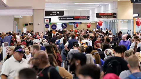 Hundreds of people stand in long queues inside an airport, signs overhead point travellers in the direction of TSA