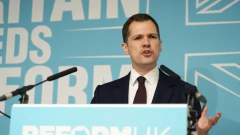 Robert Jenrick in a dark suit speaking at a lectern with light blue Reform colours