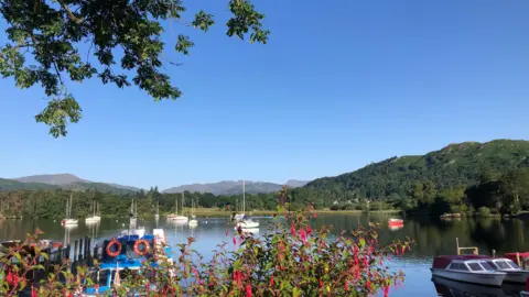 BBC Weather Watchers/ Mrs M Boat marine in Ambleside on a hot sunny day. There are pink flowers in a bush in the foreground and green hills in the background. 