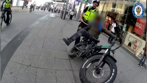 A black e-bike is stopped on a pavement with a police officer standing behind it.