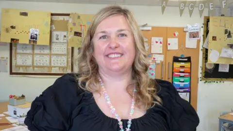 Qays Najm/BBC A woman standing in a classroom. She has curled blonde hair and is wearing a black top with a beaded long necklace around her neck. 
