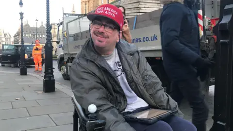 A man sits in front of Parliament in a wheelchair with a computer tablet on his lap. 