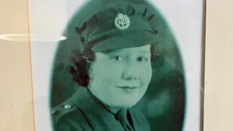 Wilkins Family An old black-and-white, framed portrait of Elsie Wilkins as a young woman. She is wearing an Auxiliary Territorial Service hat and uniform. She is looking at the camera. 