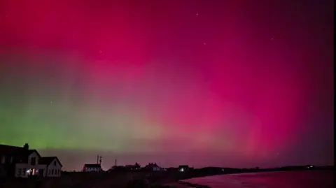 BBC weather watcher / Cara The Northern Lights lighting up the sky in pink and green colours. A number of homes are visible along a coast. 
