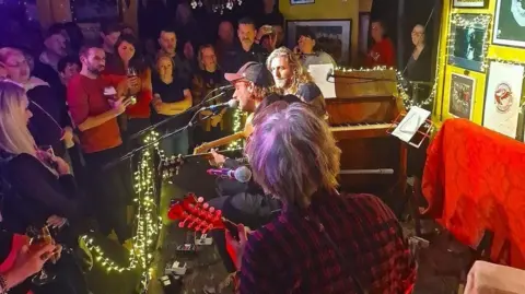 Alex Hunt Two musicians sit on a low stage playing guitars. One is wearing a black cap and another has long blonde hair. There are fairy lights over a piano and a crowd of people are standing in the audience around them. 