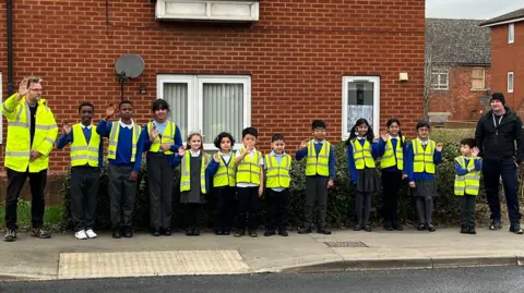 12 children line up along a pavement next to a road, all in high-vis jackets over their uniform, and are waving. There is a red brick building behind them and an adult either end of the line.